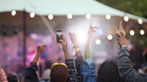 Getty Images A rear view of a crowd at a music festival. People have their arms up celebrating while one is recording on their phone. 
