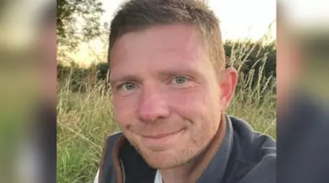 Keele University Dr Matt Clarke, who has short, dark hair and a light-coloured stubble looks at the camera, with a smile. He is wearing a grey fleece jacket, with a brown seam, and is standing in a field, with tall grass a hedge and a tree behind him.