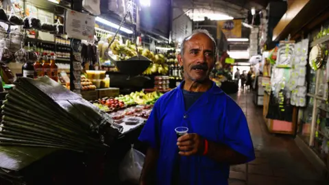 BBC A man in a Caracas market speaks while holding a glass of coffee
