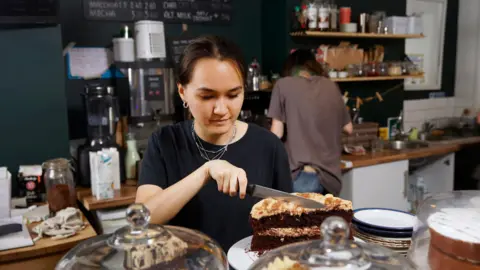 Getty Images A young woman in a cafe cutting a slice of cake with a neutral expression on her face 