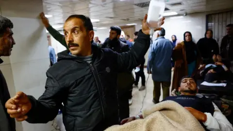 Reuters A man, looking anxious, holds a drip attached to a patient lying down in a busy hospital corridor