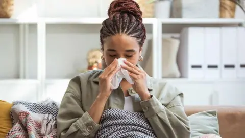 Getty Images A young woman sits on her sofa, blowing her nose into a tissue
