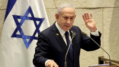 EPA Prime Minister Benjamin Netanyahu, wearing a dark suit, white shirt and navy  blue tie, with the right hand resting on a podium with two microphones, as his left arm is raised. The Israeli flag is in the background.