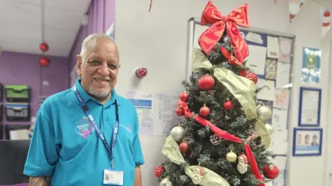 Aisha Iqbal/BBC An older gentleman, in his 70s, stands smiling in front of a beautifully adorned Christmas tree and a notice board pinned with cards. He is South Asian. He wears a blue t shirt and a lanyard, both of which identify him as an NHS volunteer. The space around him is festooned with Christmas decorations. 