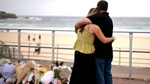 Saeed KHAN / AFP via Getty Images Mourners embrace near tributes piled together in memory of the victims of a shooting at Bondi Beach. A woman wearing a yellow top and black skirt hugs a man wearing a black polo and blue jeans as they look at a pile of flowers. In the background Bondi beach can be seen with people walking on the sand and sea in the distance