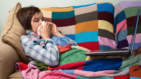 Getty Images Woman wrapped in a colourful knitted blanket lying on a sofa with a laptop on her knees. She is blowing her nose into a hankerchief.