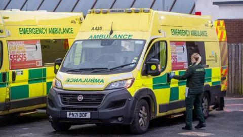 Getty Images A female paramedic opening the door to an ambulance. Another ambulance is parked alongside.