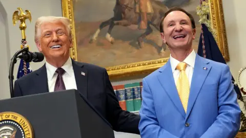 Getty Images U.S. President Donald Trump in a black suit and Louisiana Governor Jeff Landry in a blue suit smile as they stand at a podium.