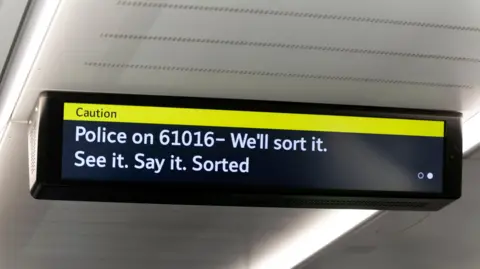 Getty Images An electronic display inside a London Underground train showing a yellow “Caution” banner and a message advising passengers to contact police on 61016, alongside the slogan “See it. Say it. Sorted”.