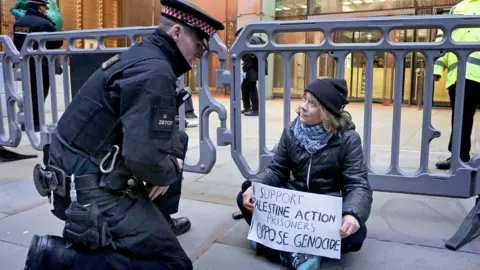 PA Media Greta Thunberg sitting on the pavement behind police barriers, holding a placard reading “I support Palestine Action prisoners. Oppose genocide,” as a police officer kneels to speak to her outside an office building in the City of London.