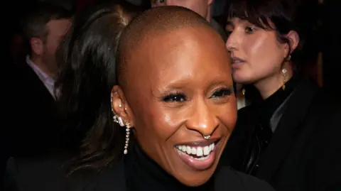 Getty Images Cynthia Erivo smiling at a Wicked: For Good event, she is wearing several diamond earrings and a diamond nose ring. Her head is shaved.