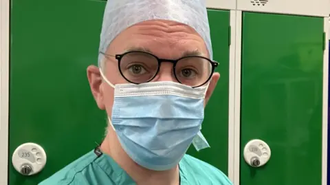 James Gallagher Head and shoulder selfie of correspondent James Gallagher, in green surgical scrubs, a blue facemask and a blue surgical hat. He is wearing dark blue glasses with metal arms. He is standing in front of a row of dark green lockers