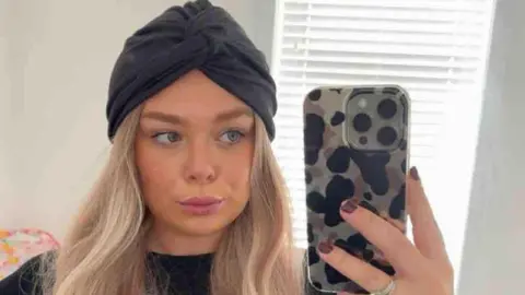 Ellie Philpot Ellie wearing a blonde wig and a black headband is taking a selfie on her iphone in the mirror. 