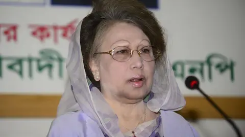 Getty Images Khaleda Zia wears a pair of wire-rimmed glasses, a light purple headscarf and a purple dress while speaking at a podium in front of a microphone
