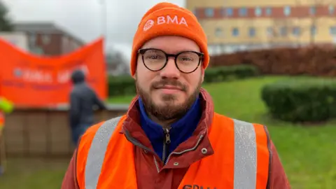 BBC A man with brown facial hair and black glasses with rain on the lenses. He is wearing an orange beanie with white writing that says "BMA". He is wearing a red waterproof jacket and an orange and silver hi-vis jacket on top.