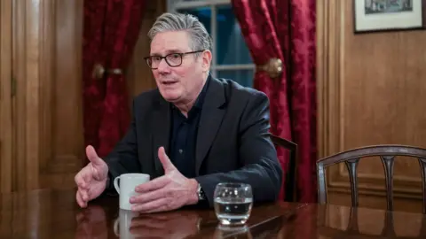 Bloomberg via Getty Images Keir Starmer gestures as he speaks while sitting in a room inside Downing Street wearing a dark shirt and suit with a mug and a glass of water on the table in front of him on Monday.