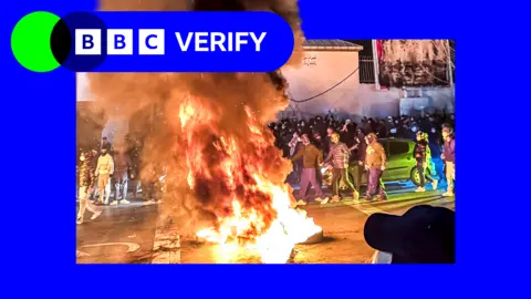 BBC Huge fire and plume of smoke in the centre of the image with crowds of masked people surrounding it during Tehran protest 