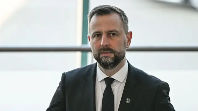 Kosiniak-Kamysz looking stern. He wears a suit, and is sliding one hand across his stomach into the suit