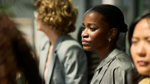 Getty Images A young black woman with her hair tied back in a bun gazing with a concerned look on her face during a meeting with other female colleagues blurred out of focus around her