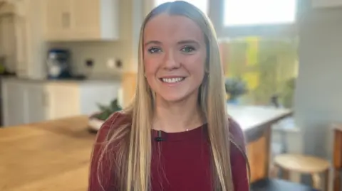 BBC Emily smiling. She has very long straight blonde hair and is wearing a burgundy top.