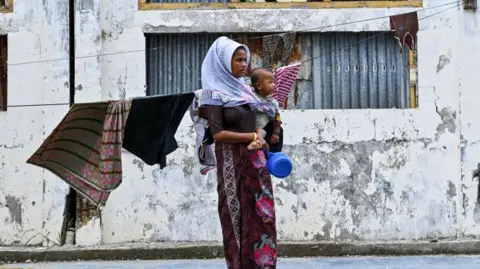 AFP via Getty Images A Rohingya refugee carries her child as she stands at a temporary shelter at a government building in Banda Aceh on December 30, 2023