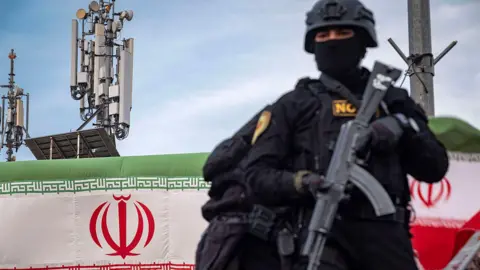 NurPhoto via Getty Images An armed Special Units police officer in Iran, dressed in black and holding a weapon, is standing on an armored vehicle while monitoring the surroundings. In the background, telecommunications towers and the Iranian flag can be seen. The photo was taken during a pro-government gathering in central Tehran.
