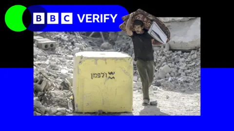 BBC A man carrying a folded mattress in Gaza walks past a yellow block placed by the Israeli military, with rubble visible in the background. The image has been imposed over the BBC Verify logo and colours. 