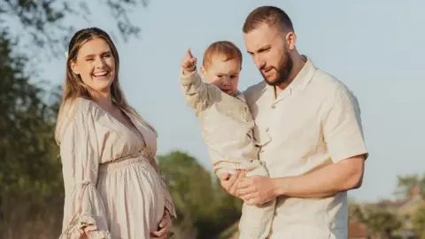 Dandelion & Rose Photography Chloe and her family all wear beige and stand outside in the sunshine with trees visible in the background as part of a professional family photoshoot. Chloe is visibly pregnant and holds her stomach smiling, while her partner holds their son.
