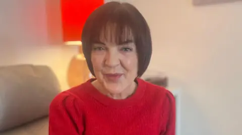 BBC Jennifer has brown hair which is styled in a bob and a fringe. She is sitting on the sofa wearing a red round-neck top. Behind her is a lamp which has a red lampshade. She is wearing makeup.