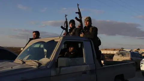 AFP Syrian government forces hold rifles while riding on a pick-up truck on the road to Raqqa, Syria (19 January 2026)